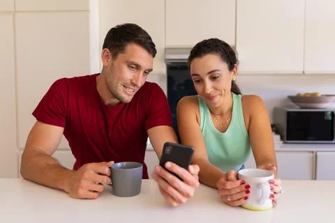 Caucasian couple using smartphone while having coffee in the kitchen at home 写真素材