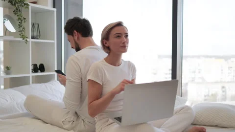 Caucasian coupleworking on gadgets while sitting back to back on bed. Stock Footage 255112185