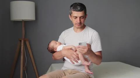 Caucasian father currently using the mobile phone while holding newborn baby on 스톡 동영상 291309000