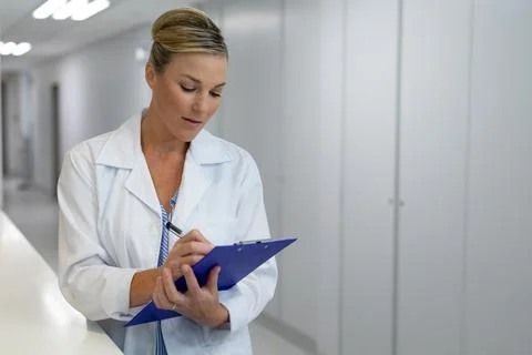 Caucasian female doctor standing in hospital corridor filling medical chart Stock Photos