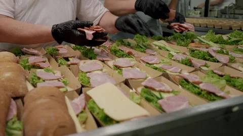Caucasian female worker in gloves assembles sandwiches with lettuce, ham, and Stock Footage 340172581