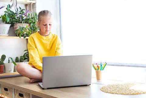 Caucasian girl sitting on the windowsill, near houseplants in front of a laptop. Stock Photos