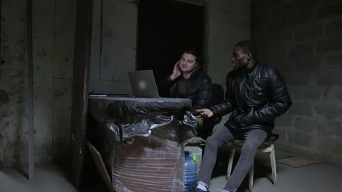 Caucasian guy and black guy are sitting near open laptop in a basement during a Stock Footage 171348506