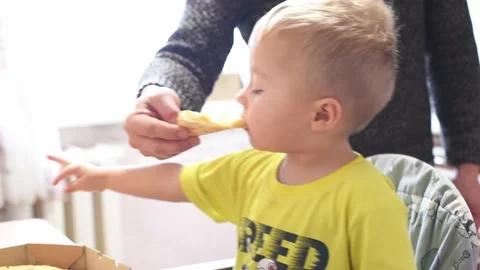 Caucasian kid boy sits in the kitchen and eats pizza. Father feeds his son with Stock Footage 221360024