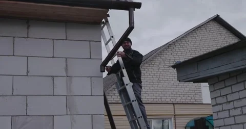 A Caucasian man attaches a gutter. A lone man climbs a ladder to attach a Stock Footage 323570993