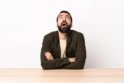 Caucasian man with beard in a table looking up and with surprised expression Stock Photos