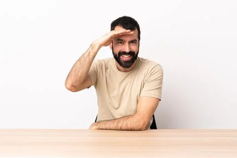 Caucasian man with beard in a table looking far away with hand to look someth Stock Photos