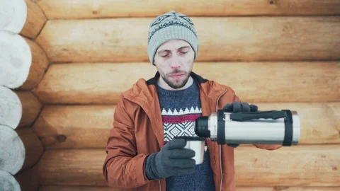 A Caucasian man in a brown jacket, hat and gloves drinking hot tea pouring it Stock Footage 128124365