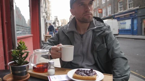 Caucasian Man Drinking Tea with Crumpets In London Wearing Rain Jacket and Hat Stock Footage 89700479