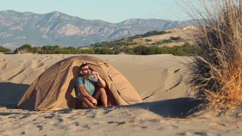 Caucasian man drinks water while sitting in a tent in the sand dunes at sunrise  Stock Footage 118145097