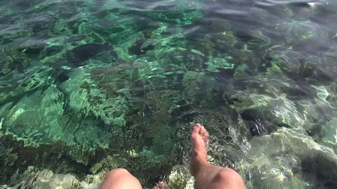 Caucasian man sits and splashes his bare feet in azure transparent sea. POV shot Stock Footage 76989595