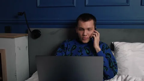 Caucasian man talking on the phone while working with a laptop on bed at home 스톡 동영상 144629954