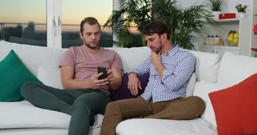 Caucasian Men Sitting on the Sofa Talking and Surfing Internet on Mobile Phone Stock Footage 85483196