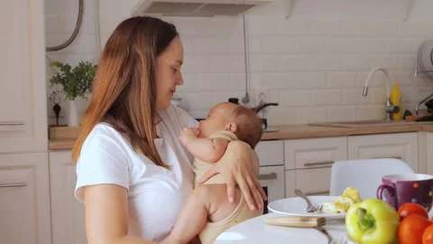 Caucasian mom rocking newborn while eating lunch at kitchen table comforting and Видео 291281499