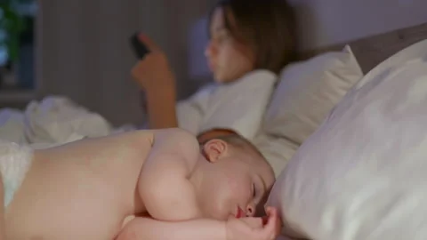 Caucasian mother using smartphone while baby sleeps peacefully beside her on a Stockbeeldmateriaal 341524129