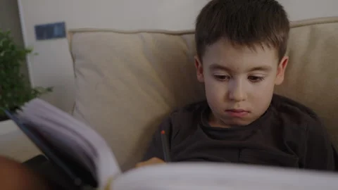 Caucasian schoolboy sitting on sofa doing homework, holding pen to write. Boy Stock Footage 253842861