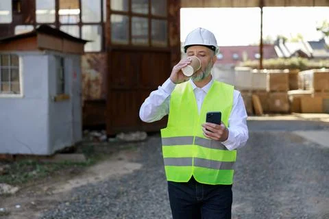 Caucasian senior foreman browsing internet on modern smartphone and drinking hot Stock Photos
