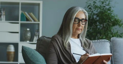 Caucasian senior lady in the glasses sitting on the couch and writing down Video stock 103701341