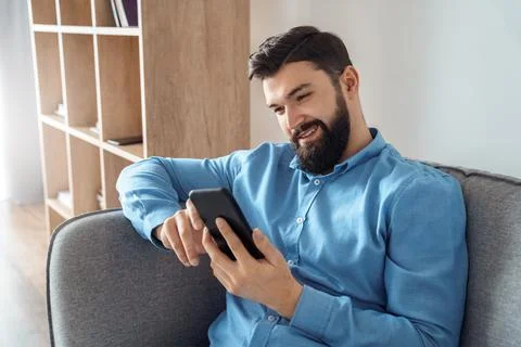 Caucasian smiling man using smartphone device relaxing sitting on home sofa Stock Photos