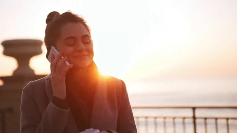 Caucasian woman speaking mobile phone and laughing during sunrise at sea in Stock Footage