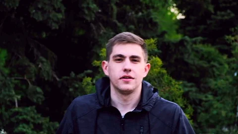 Caucasian young guy in a black jacket walks in the park and talks to the camera Stock Footage 129688401