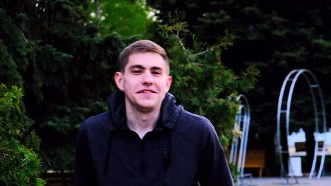 Caucasian young guy in a black jacket walks in the park and talks to the camera Stock Footage 129688422