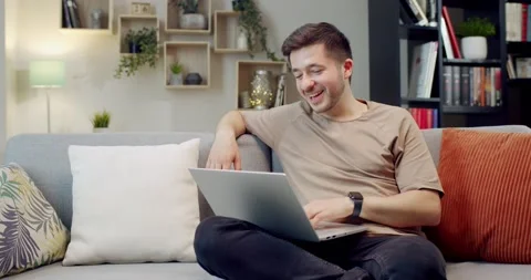 Caucasian young smiling man sitting on cozy couch at home and working on laptop Stock Footage 220369953