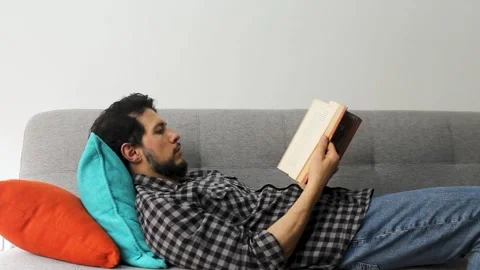A caucasic latin man lying on a couch in a relaxing attitude leafing a text book Stock-Footage 340583329