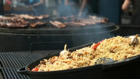 Cauldron with steaming pilaf in the foreground. Large electric rotating grill Stock Footage 129336744