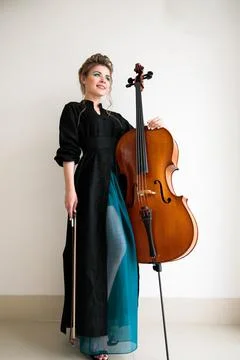 Cellist posing while standing with a cello against a light wall Stock Photos