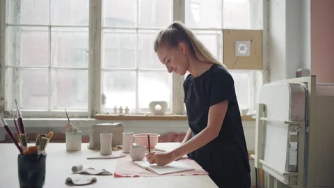 Ceramic artist looking at clay mug and sketching design Stock Footage 106829486