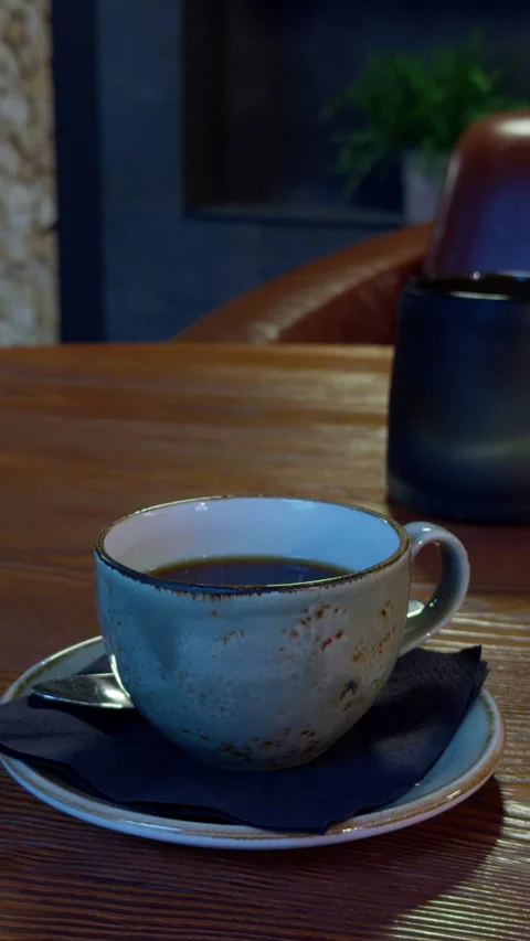Ceramic coffee cup on saucer with spoon and napkin on wooden table in cozy cafe Stock Footage 345765866