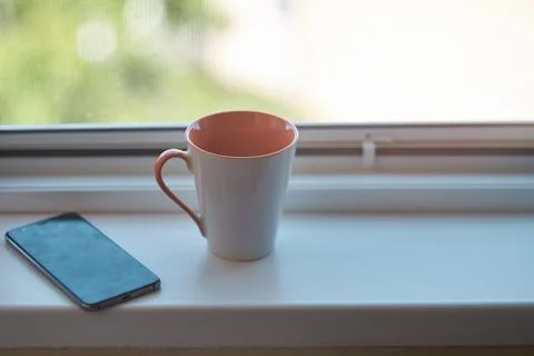 Ceramic coffee mug and a modern smartphone resting on a bright window sill .. Stock Photos