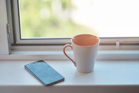 Ceramic coffee mug and smartphone resting on a white window sill at home Stock Photos