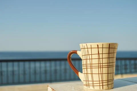 A ceramic coffee mug sitting on a table with ocean view in the background Stock Photos