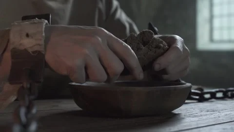 Chained hands of a prisoner snapping a rye loaf over a bowl inside a gloomy cell Stock Footage 338991632