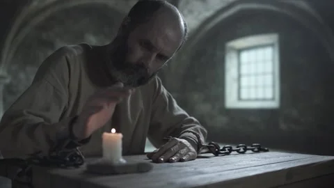 Chained prisoner looking at a burning candle and warming his hand over a flame Vídeo Stock 339232484