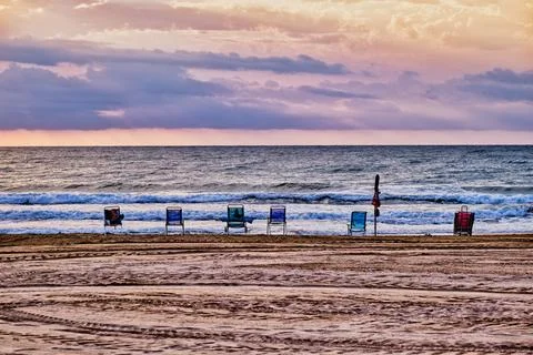 Chairs lined up on sandy beach facing ocean waves during sunset near coastlin Stock Photos
