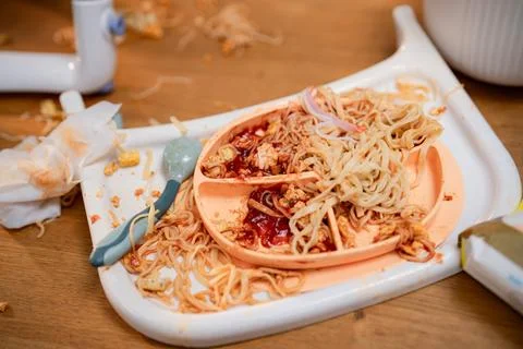 A chaotic dining table shows an orange plate filled with noodles and sauce. Foto stock