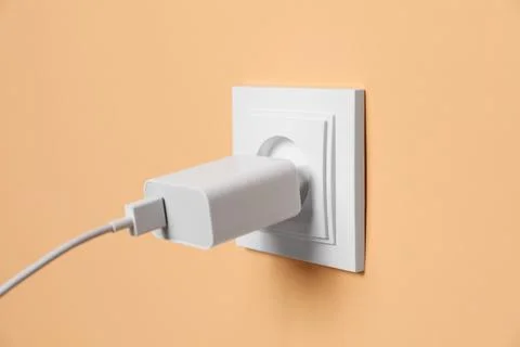 Charger adapter plugged into power socket on pale orange wall, closeup. Ele.. Stock Photos