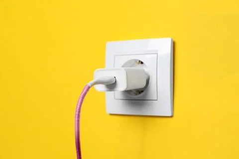 Charger adapter plugged into power socket on yellow wall, closeup. Electric.. Stock Photos
