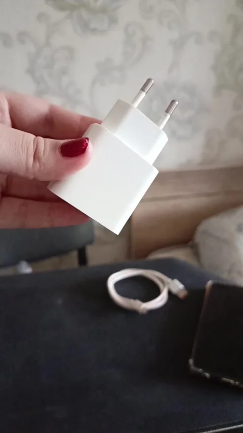A charging unit in a womans hand, a cable and a phone on the table. Phone Stock Footage 319980596