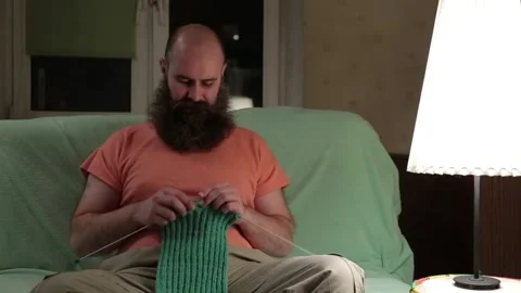 A charismatic man with a beard knits a scarf while sitting in a room on couch Stock-Footage 87829890