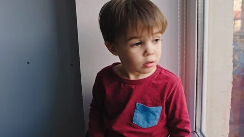 Charismatic small cute boy looking sad through the window he stay at home in Stock Footage 127920267