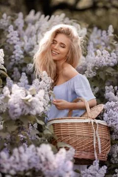 Charming blonde model against the backdrop of lilac bushes holds a straw basket Stock Photos
