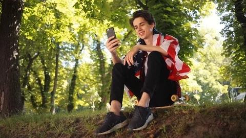 Charming boy with American flag using mobile phone smile sitting on skate in Stock Footage 92048306