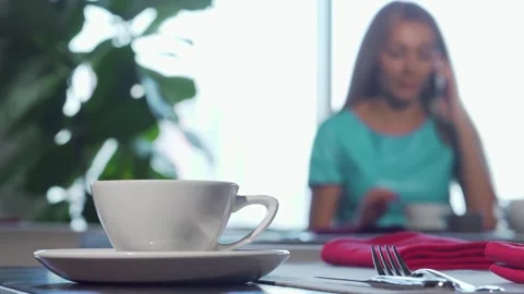 Charming cafe scene with a cup in focus and a woman enjoying a phone call Stock Footage 311185911