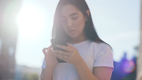 Charming casual brunette actively using modern smartphone outdoors in bright Video stock 111219911