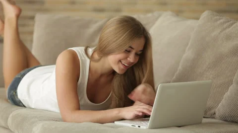 Charming girl lying on sofa using laptop looking at camera and smiling. Panning Stock Footage 34114841