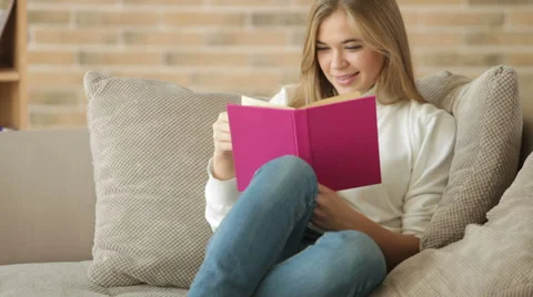 Charming girl sitting on sofa reading book closing it looking at camera  Stock Footage 34114520